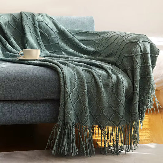 Navy blue knitted throw blanket with an elegant diamond pattern and fringe detailing draped over a blue armchair in a cosy living room setting.