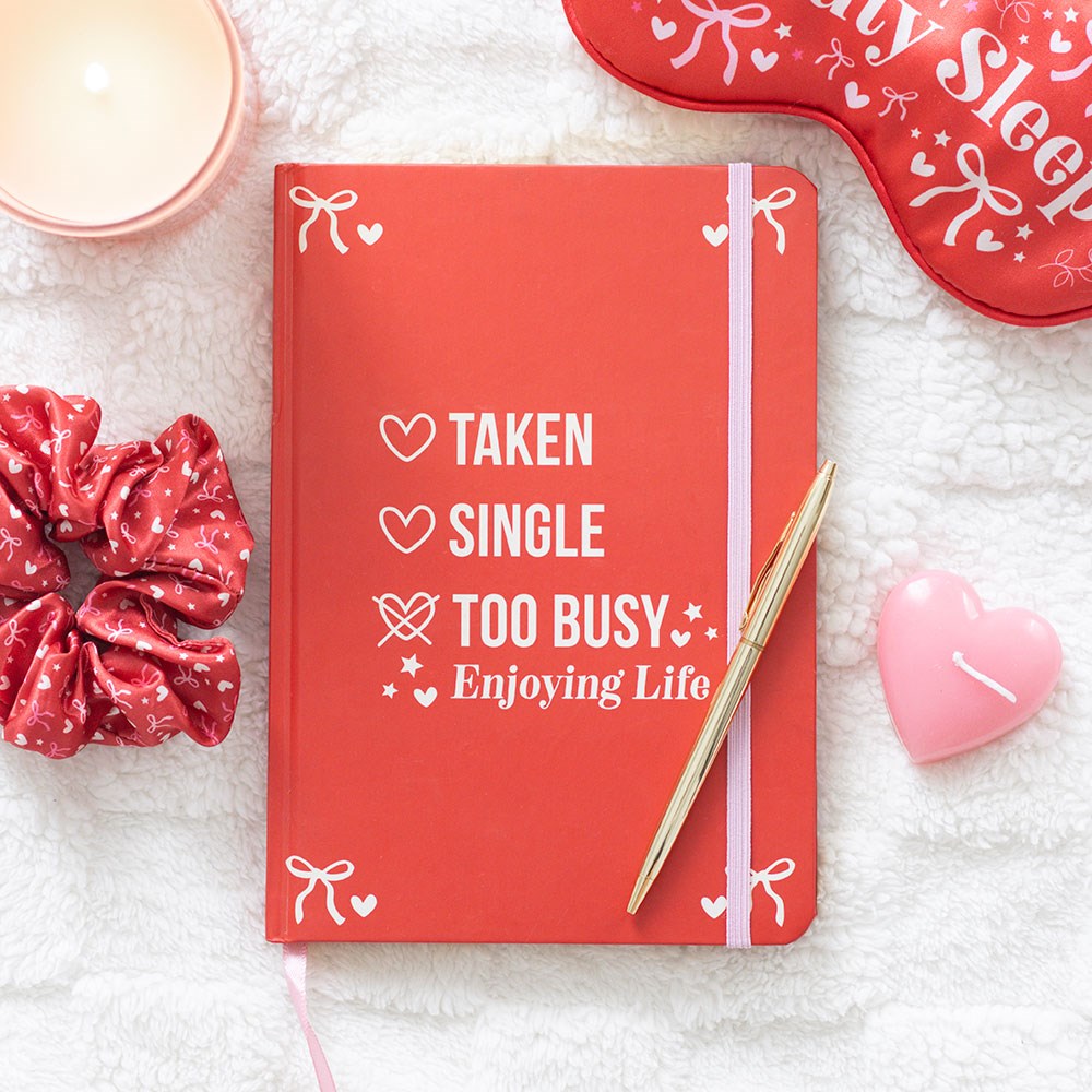 Red notebook with “Taken, Single, Too Busy Enjoying Life” text, displayed with a pen and cozy accessories on a soft background.