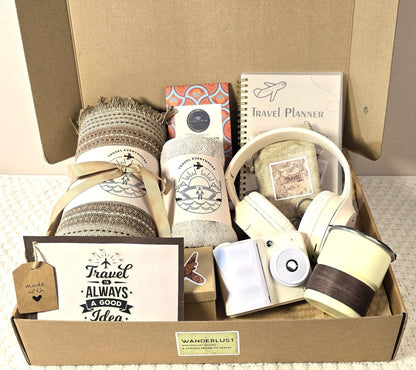 Travel-themed gift box featuring a cozy blanket, soft towel, stylish headphones, instant camera, travel planner notebook, reusable cup, chocolate bar, and travel stickers — beautifully packed and perfect for adventurers and travelers.