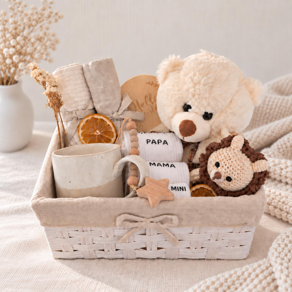 A baby gift basket set in a white woven basket, filled with a cream teddy bear, a crocheted lion plush, a ceramic mug, folded beige textiles, and dried orange slices, styled on a soft cream fabric with a vase of dried flowers in the background.