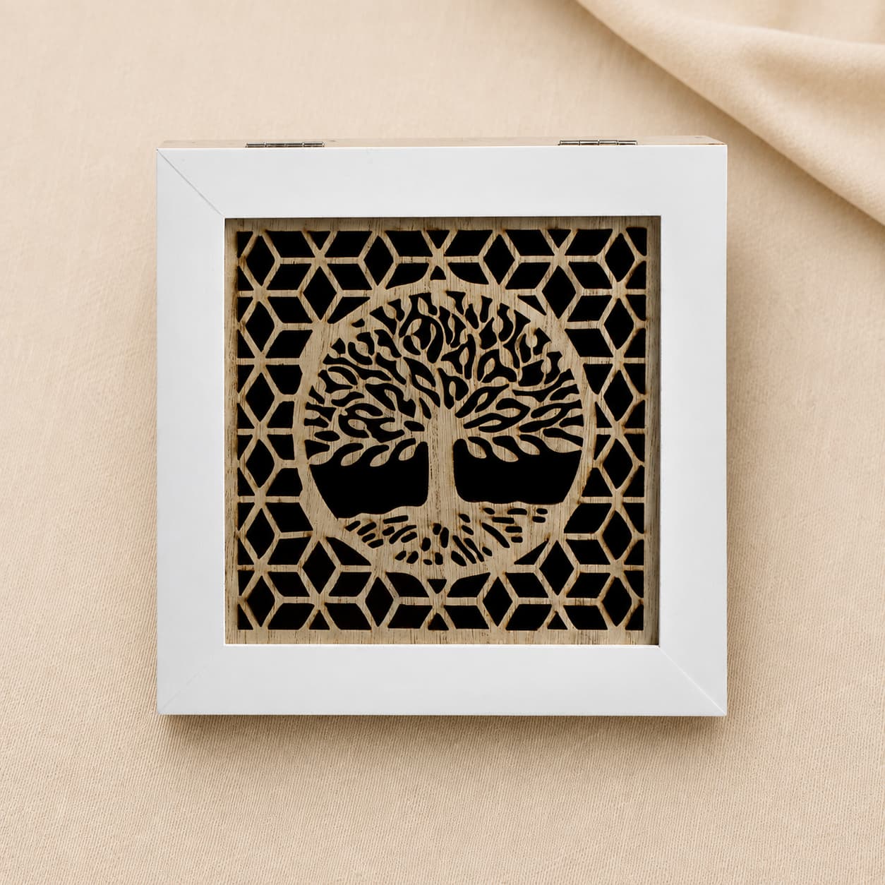 A square wooden babies keepsake box with a light natural wood grain body and a white-trimmed lid featuring a laser-cut gold tree of life design over a black geometric background, set on a beige fabric surface.