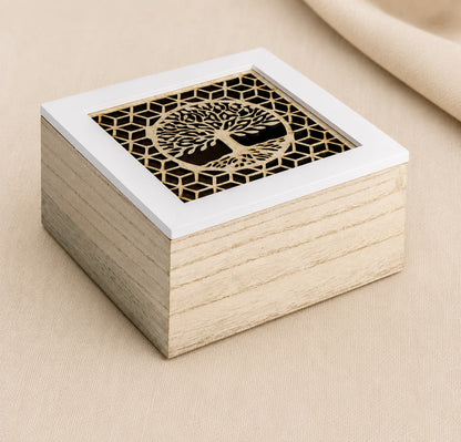 A square wooden babies keepsake box with a light natural wood grain body and a white-trimmed lid featuring a laser-cut gold tree of life design over a black geometric background, set on a beige fabric surface.