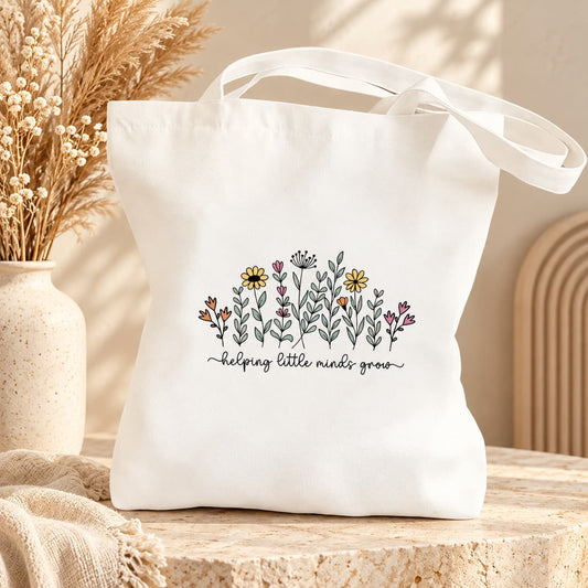 White floral teacher tote bag with "helping little minds grow" design, styled with dried flowers against a neutral boho background