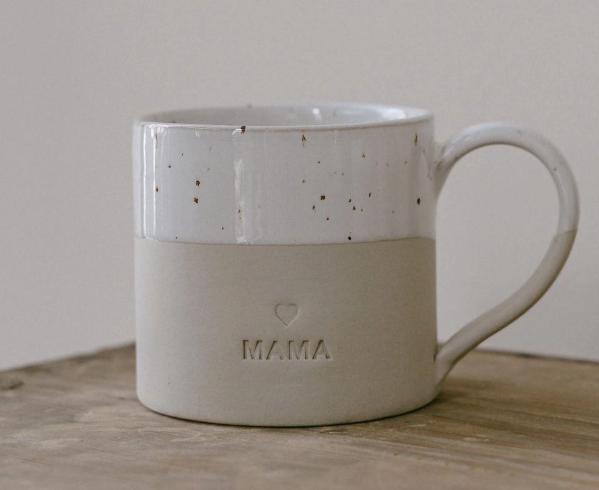 A ceramic mug with a speckled white upper half and a matte beige lower half embossed with the word 'MAMA' and a small heart, resting on a wooden surface against a neutral background.