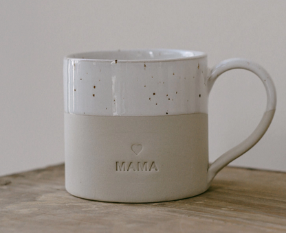 A ceramic mug with a speckled white upper half and a matte beige lower half embossed with the word 'MAMA' and a small heart, resting on a wooden surface against a neutral background.