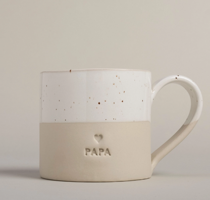 A ceramic mug with a two-tone design, featuring a white upper half with brown speckles and a matte beige lower half embossed with the word 'PAPA' and a small heart.