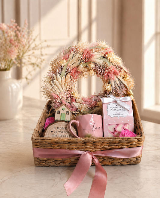 Luxury gift basket featuring a dried floral wreath, pink ceramic mug, wooden house keepsake, marshmallow gift box, and decorative home plaque arranged in a woven basket with a pink ribbon.