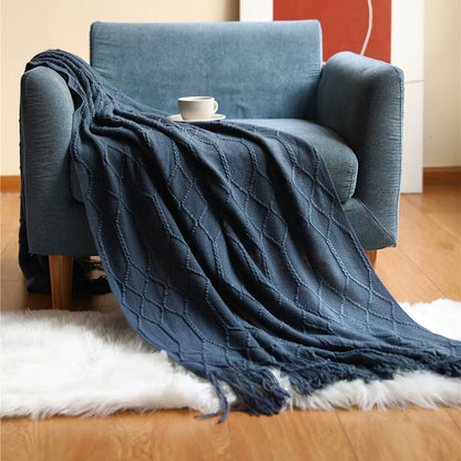 Navy blue knitted throw blanket with an elegant diamond pattern and fringe detailing draped over a blue armchair in a cosy living room setting.