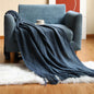 Navy blue knitted throw blanket with an elegant diamond pattern and fringe detailing draped over a blue armchair in a cosy living room setting.