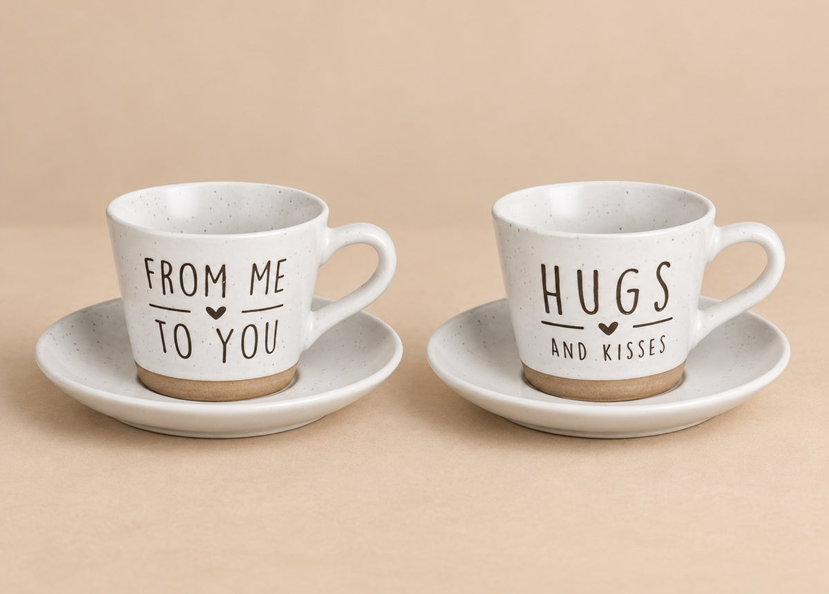 Two speckled white ceramic espresso cups with saucers on a beige background. One cup is printed with 'FROM ME TO YOU' and the other with 'HUGS AND KISSES' in dark brown lettering.