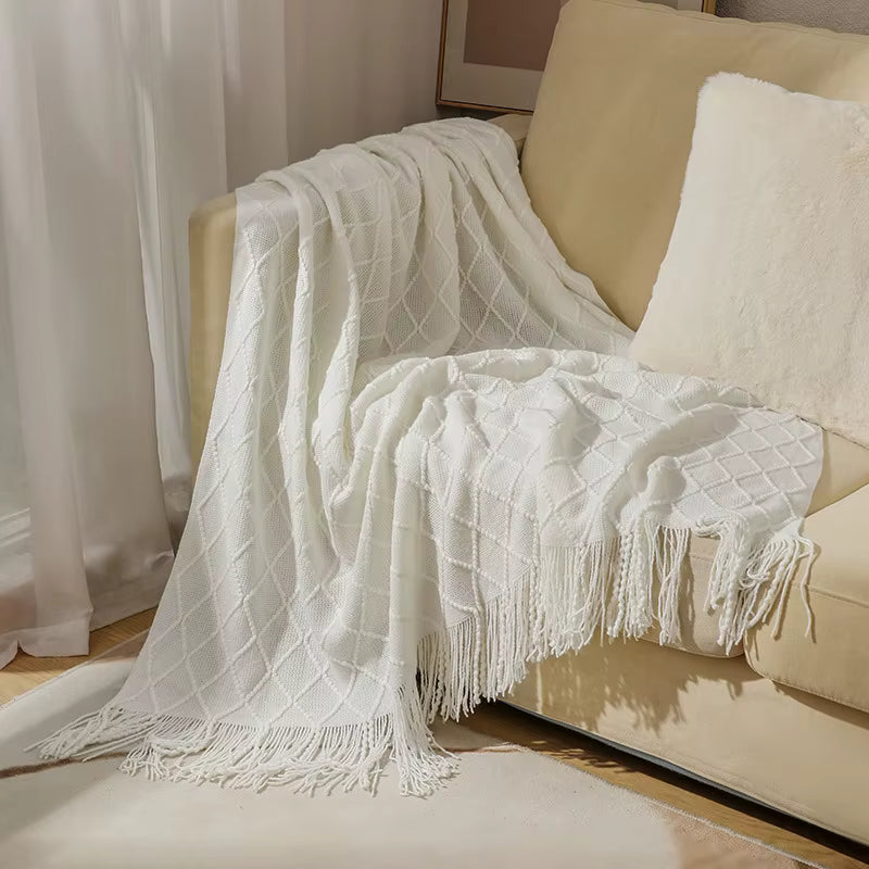Ivory white knitted throw blanket with a textured diamond pattern and decorative fringe draped over a cream sofa in an elegant living room setting.