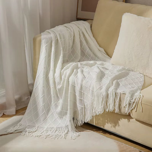 Ivory white knitted throw blanket with a textured diamond pattern and decorative fringe draped over a cream sofa in an elegant living room setting.