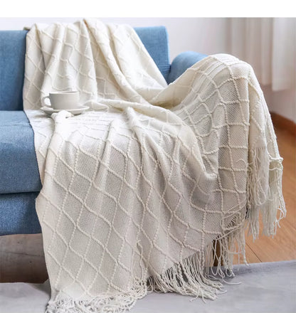 Ivory white knitted throw blanket with a textured diamond pattern and decorative fringe draped over a cream sofa in an elegant living room setting.