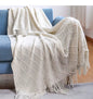 Ivory white knitted throw blanket with a textured diamond pattern and decorative fringe draped over a cream sofa in an elegant living room setting.