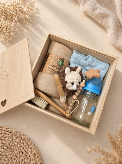 Open wooden newborn baby keepsake gift box with muslin blanket, 100% cotton onesie, glass baby bottle, rattle, pacifier holder, and newborn hair brush in a warm neutral boho setting.