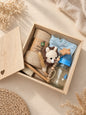 Open wooden newborn baby keepsake gift box with muslin blanket, 100% cotton onesie, glass baby bottle, rattle, pacifier holder, and newborn hair brush in a warm neutral boho setting.