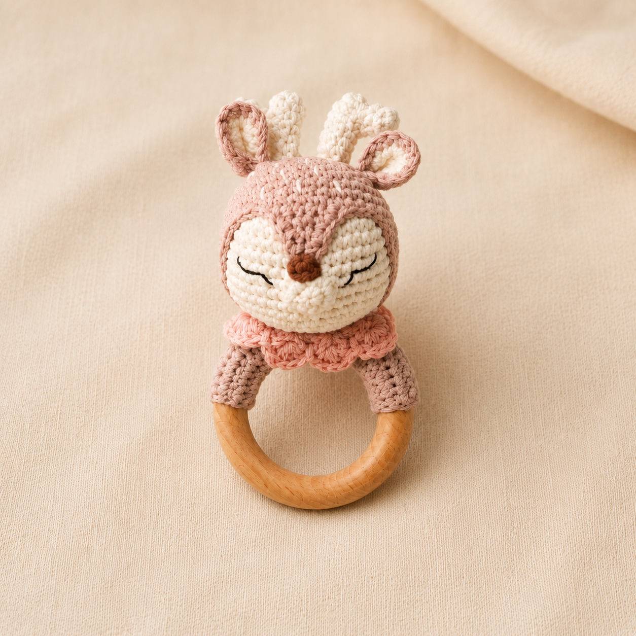 Pink and cream crochet deer baby rattle with antlers and natural wooden teething ring on beige fabric.
