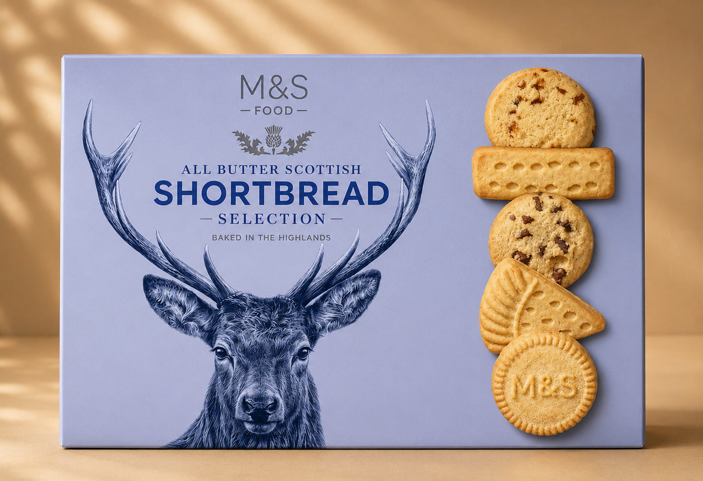 M&S Food All Butter Scottish Shortbread Selection box, featuring a light blue background with a dark blue deer illustration and a stack of four golden-brown shortbread cookies on the right side.