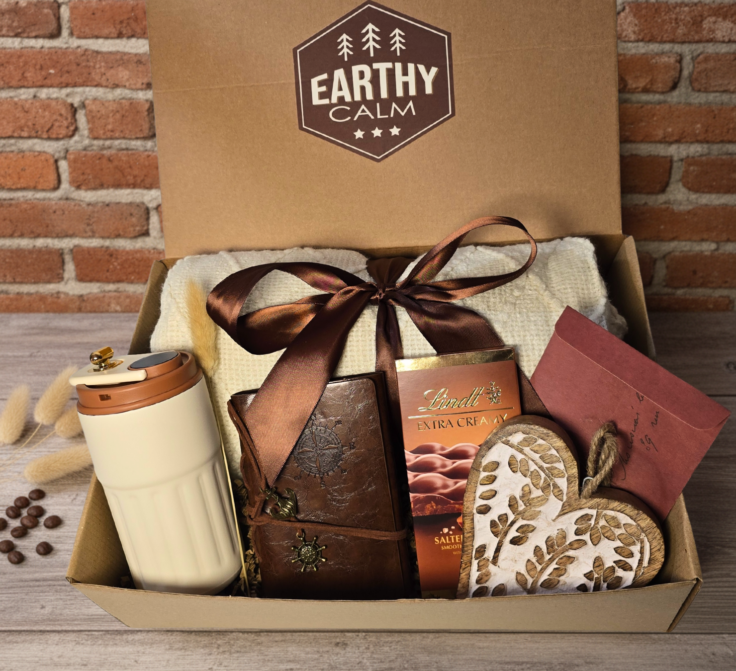 Earthy Calm rustic gift box featuring a leather-look journal with compass charm, cream travel mug, knitted blanket, Lindt chocolate, and wooden heart accessory.