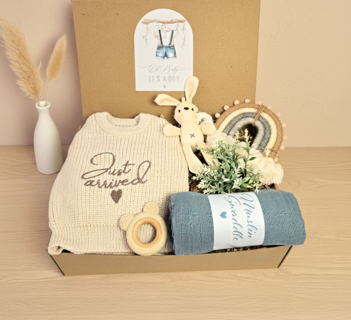 New Baby Boy gift box featuring a 'Just Arrived' knitted sweater, blue muslin swaddle, wooden teether, plush bunny toy, and macrame rainbow decoration.