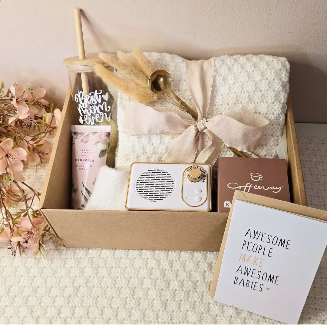“A beautifully arranged ‘Best Mom Ever’ gift box containing a cozy cream blanket tied with a ribbon, floral hand cream, reusable tumbler, gold coffee spoon, mini speaker, Coffeeway coffee box, and a greeting card that says ‘Awesome people make awesome babies,’ surrounded by soft pink flowers