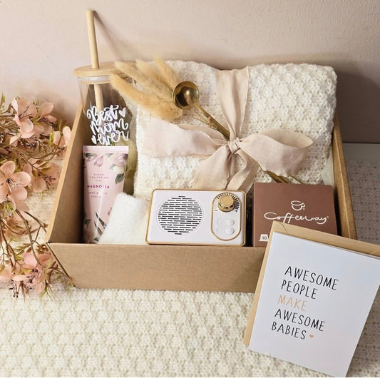 “A beautifully arranged ‘Best Mom Ever’ gift box containing a cozy cream blanket tied with a ribbon, floral hand cream, reusable tumbler, gold coffee spoon, mini speaker, Coffeeway coffee box, and a greeting card that says ‘Awesome people make awesome babies,’ surrounded by soft pink flowers
