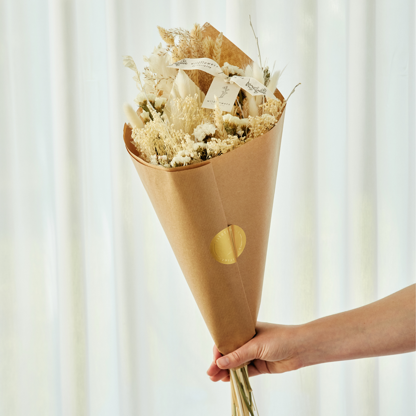 Bouquet of dried flowers wrapped in brown paper with a gold circle, held against a white curtain background.