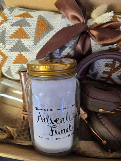 Travel-themed gift box with a large boho blanket, adventure savings jar, leather notebook, brown headset, ceramic mug, and luxury chocolate arranged in a cozy adventure-inspired setting.