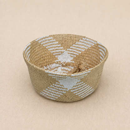 Handwoven natural seagrass baby basket with white geometric detailing on a soft ivory linen background.