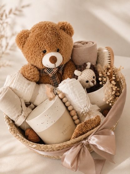 Minimal boho baby gift basket with brown teddy bear, neutral muslin cloths, ceramic baby keepsakes, wooden brush, crochet toy, dried flowers and blush ribbon.