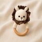 Cream and brown crochet lion baby rattle with natural wooden teething ring on beige fabric.