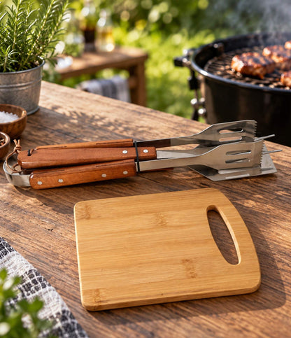A bamboo cutting board with a handle cutout sits on a wooden outdoor table next to metal grilling tools with brown wooden handles, with a grill and plants blurred in the background.