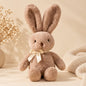 Soft brown plush bunny with long ears and cream satin bow styled on a warm beige boho-minimal background.