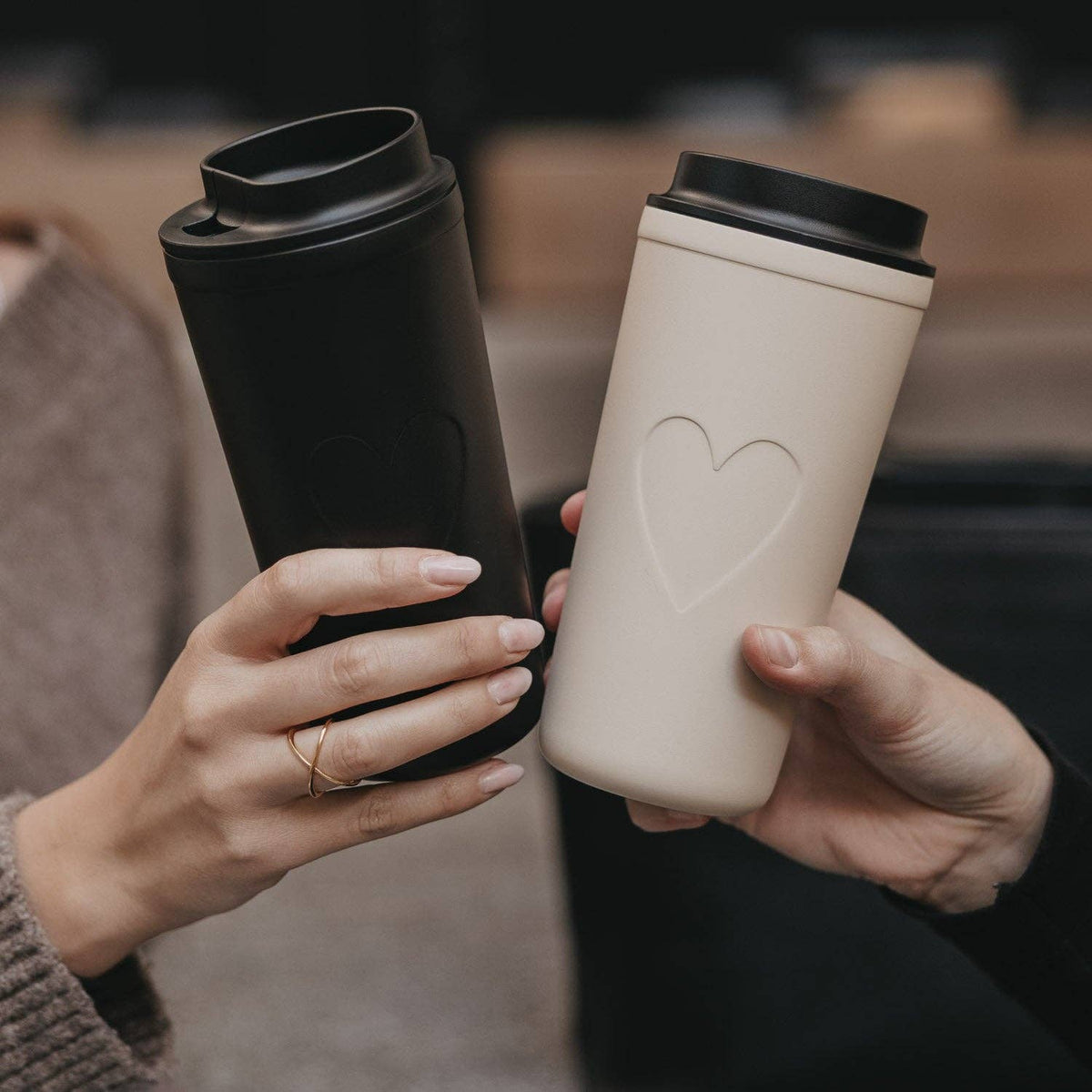 Cream heart travel cup beside a black travel cup, both held in hands.
