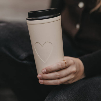 Cream 540 ml travel cup with an embossed heart and black lid, held in one hand.