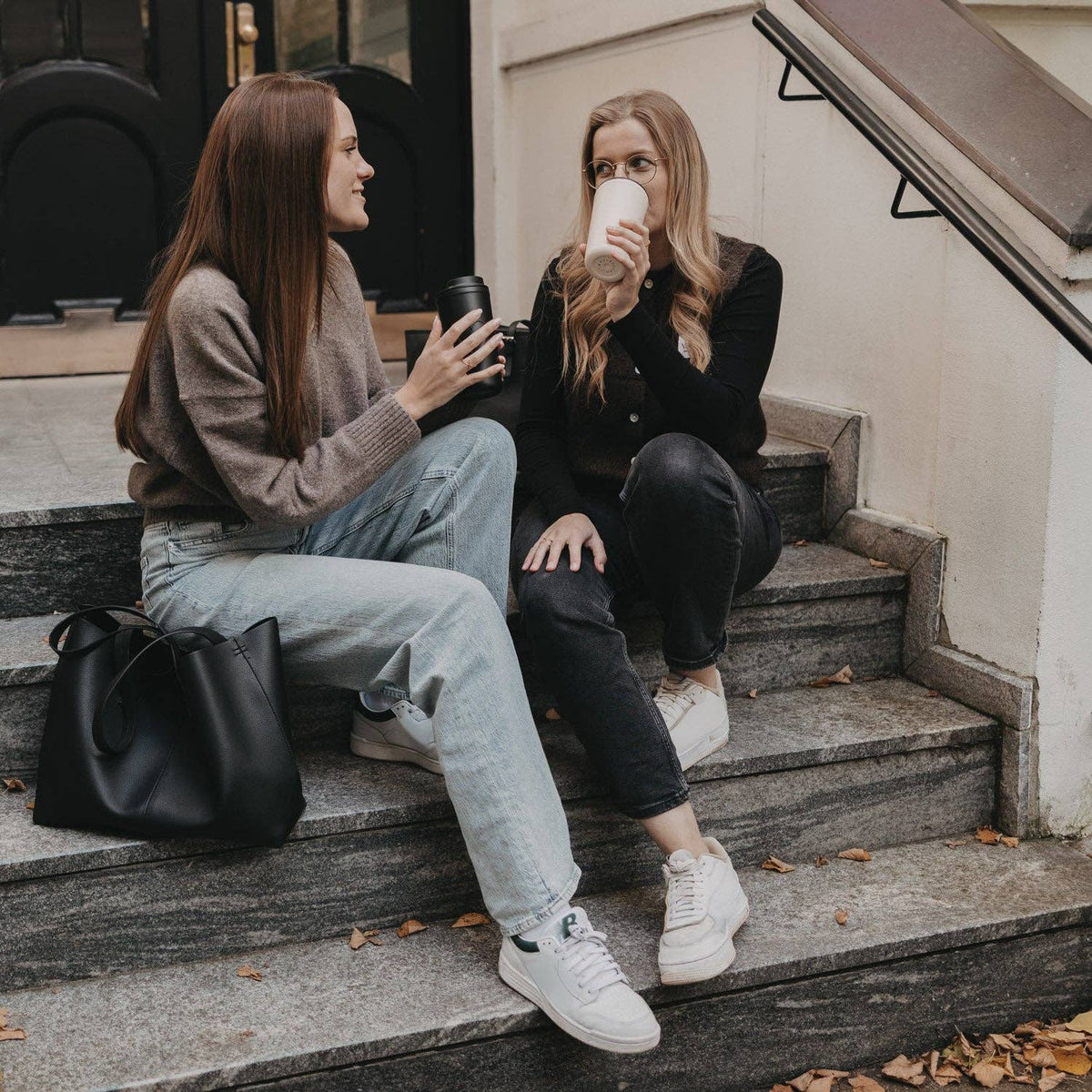 Two people sitting on outdoor steps enjoying drinks from cream and black travel cups.