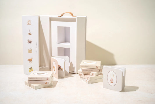Luxury newborn baby girl gift box with educational mini library books, Mustela baby cleansing gel, FRIGG pacifier, pink knitted blanket, and soft nursery-themed packaging.