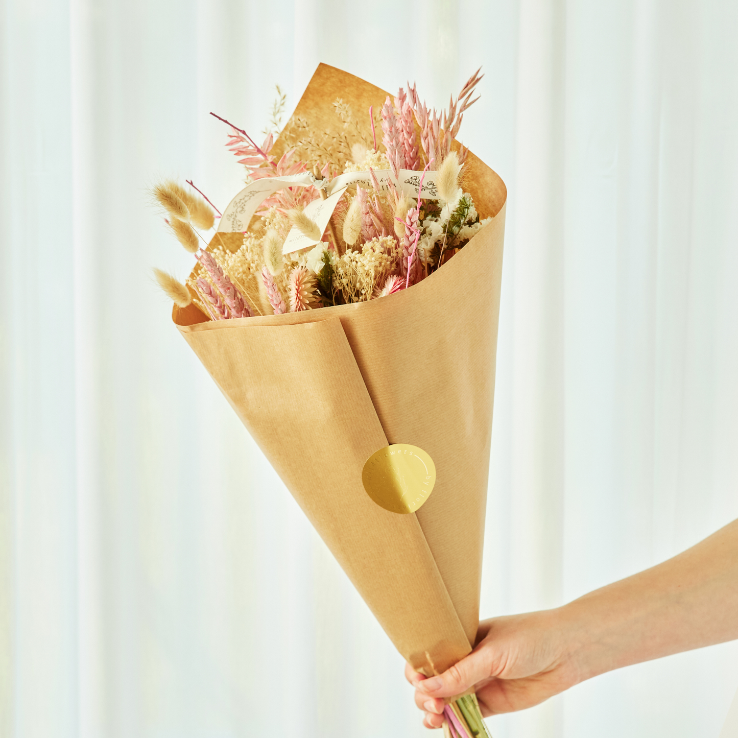 Bouquet of dried flowers wrapped in brown paper held by a hand against a white background