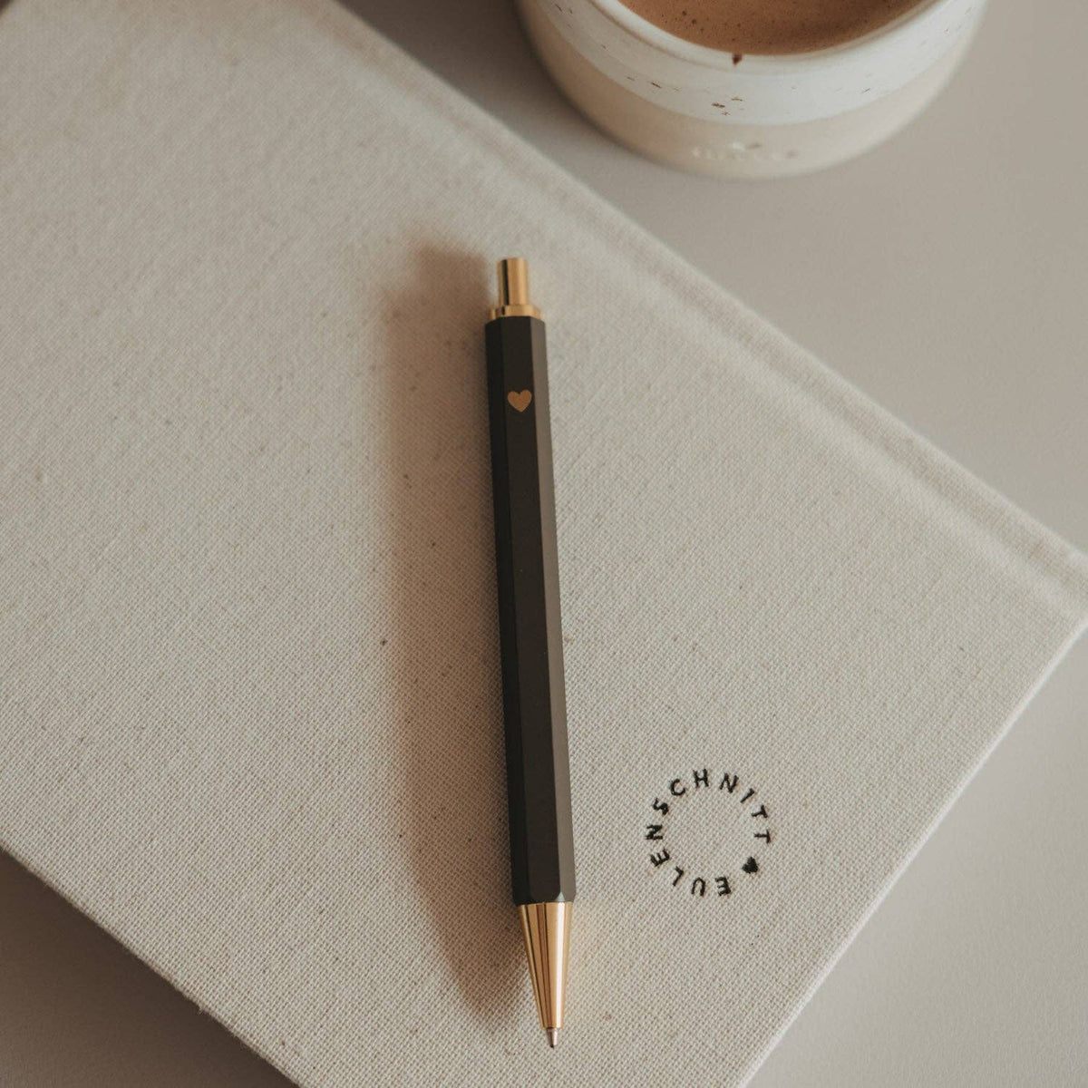 Brown Eulenschnitt ballpoint pen with a gold-coloured heart motif resting on a notebook beside a mug.