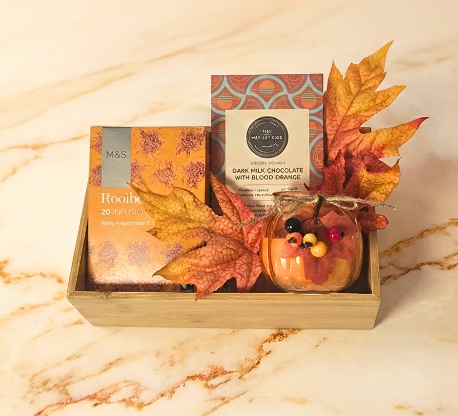 M&S Autumn Delights Gourmet Tray featuring M&S Rooibos Tea, M&S Dark Milk Chocolate with Blood Orange, and a small autumn-themed candle in a wooden tray.