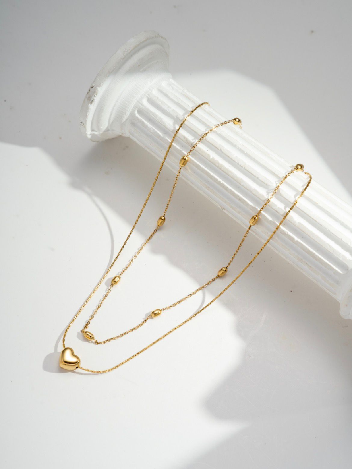 Layered gold necklace with delicate chains, small bead accents, and a heart pendant displayed on a white decorative stand.