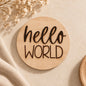 Round wooden “Hello World” newborn announcement sign styled on a warm neutral boho-minimal background.