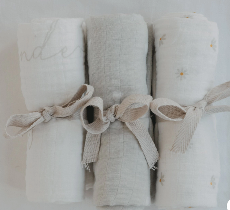 Three rolled white muslin baby swabs tied with beige ribbon bows, arranged side by side on a white background. The left swab has a gray script word, the center is plain, and the right has small daisy prints.