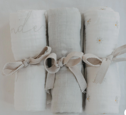 Three rolled white muslin baby swabs tied with beige ribbon bows, arranged side by side on a white background. The left swab has a gray script word, the center is plain, and the right has small daisy prints.