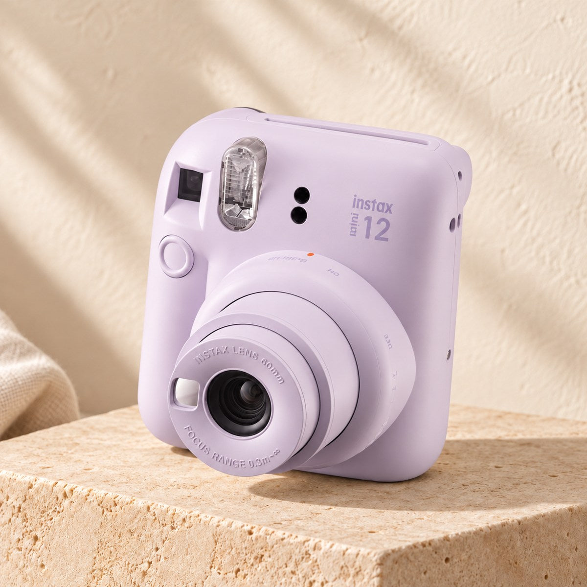 Fujifilm Instax Mini 12 in lilac purple on a beige stone display with an ivory boho background.