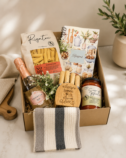 Italian-themed gourmet gift box featuring pasta, pasta sauce, rosé bottle, wooden cheese board with utensils, recipe notebook, and striped kitchen towel beautifully arranged in a kraft gift box on a neutral kitchen-inspired background.