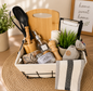 Kitchen-themed gift basket featuring a wooden cutting board, cooking utensils, wooden pestle and mortar, glass soap dispenser, salt and pepper jars, pepper grinder, decorative plant, kitchen towels, and home accessories beautifully arranged in a fabric-lined wire basket.