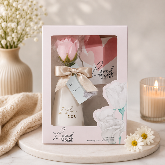 Elegant floral gift set in a blush pink presentation box featuring a ceramic vase with a pink soap rose, decorative greeting card, satin ribbon, and floral accents, beautifully displayed on a soft pink background with daisies and decorative candles.