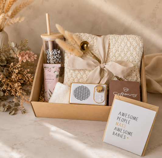 Luxury new mom gift box beautifully arranged on a marble surface, featuring a “Best Mom Ever” tumbler with bamboo lid and straw, cozy knitted blanket tied with a ribbon, floral hand cream, soft washcloth, decorative pampas grass, portable speaker, coffee sachets, and a greeting card, styled with neutral-toned flowers and elegant beige décor.