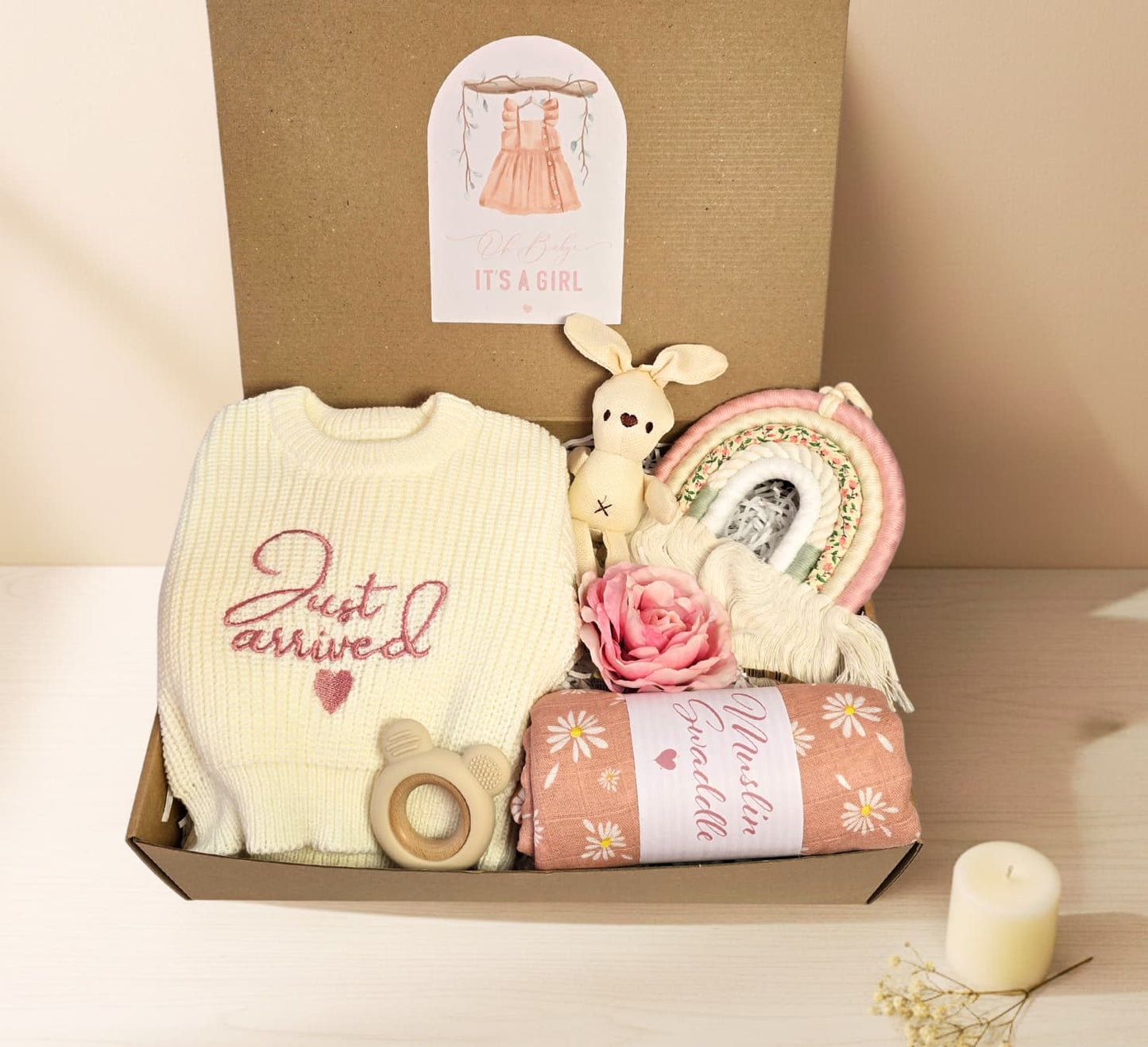 Newborn Baby Girl gift set featuring a 'Just Arrived' knitted sweater, pink daisy muslin swaddle, wood teether, plush bunny toy, and macrame rainbow.